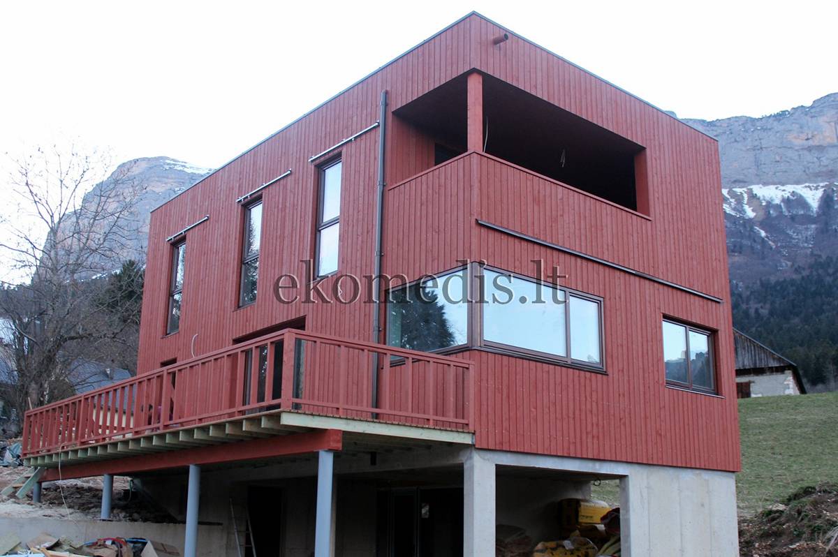 Kubeformet hus med rød stående kledning, innfelt balkong og terrasse på stolper, fjell i bakgrunnen