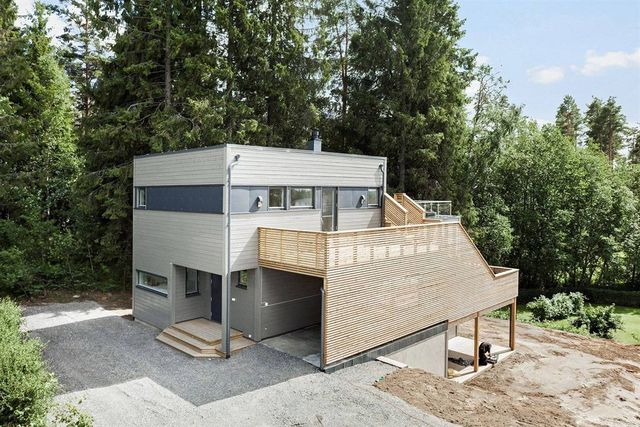 Grått trehus med flatt tak og stor treterrasse over en overbygd plass, omgitt av skog