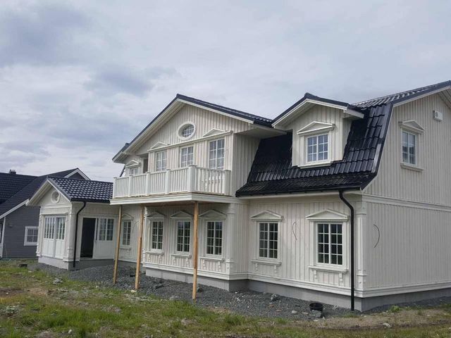 Hvitt toetasjes hus med stående kledning, svart takstein, arker og balkong under oppføring