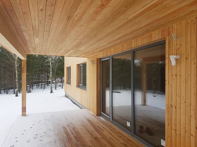 Overbygd terrasse med tregulv og panelt tak ved store skyvedører i hus med stående trekledning, snø ute