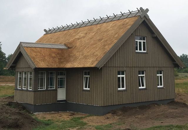 Hus med brun stående trekledning, stråtak, hvite sprossevinduer og glassveranda på nyopparbeidet tomt