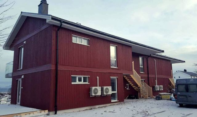 Rødt toetasjes trehus med stående kledning, utvendige tretrapper og varmepumper på fasaden, snø på bakken