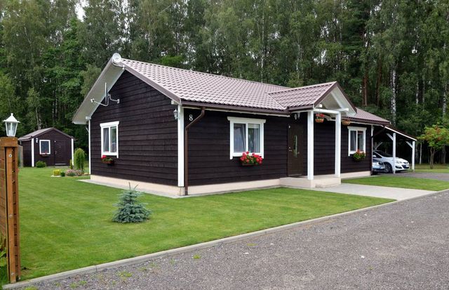 Mørkbrunt enetasjes trehus med hvite detaljer, saltak, carport og bod på plen foran skog