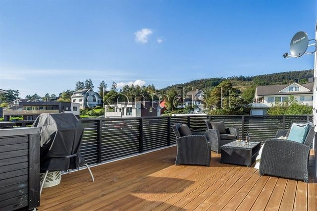 Stor terrasse med tregulv, mørkt rekkverk, utemøbler og grill, med utsikt over nabolag og åser