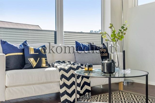 Hvit sofa med mørkeblå puter, rundt glassbord og store vinduer med plisségardiner