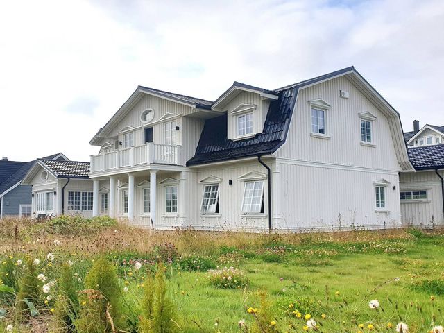 Stort hvitt trehus med stående kledning, mansardtak, søyler og balkong, sett fra en eng med markblomster