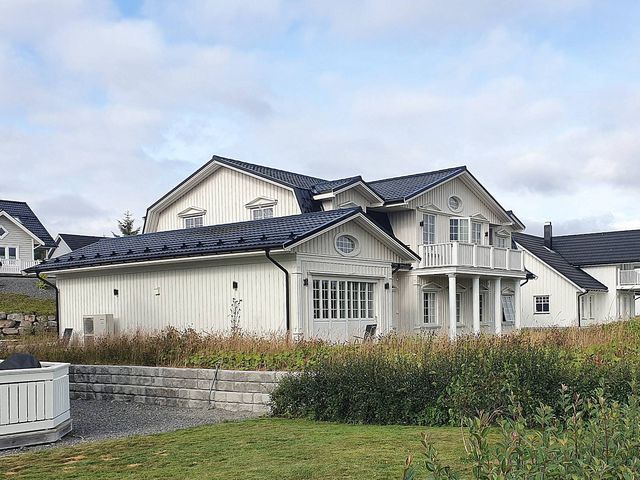 Hvitt trehus med sort takstein, sprossevinduer, søyler og balkong, med støttemur og plen foran