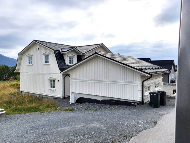 Hvitt trehus med mansardtak og tilbygg sett fra siden, gruset tomt og søppeldunker foran