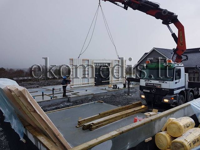 Kranbil løfter veggelement med vindu inn på betongfundament, trelast og isolasjonsruller i forgrunnen