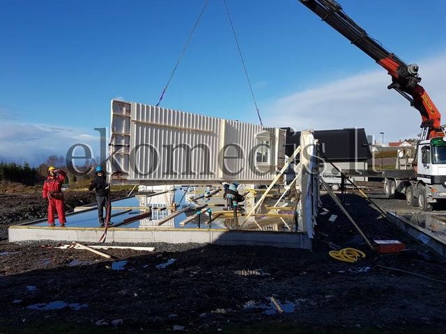 Kran løfter et veggelement med kledning på plass på betongfundamentet mens montører følger med