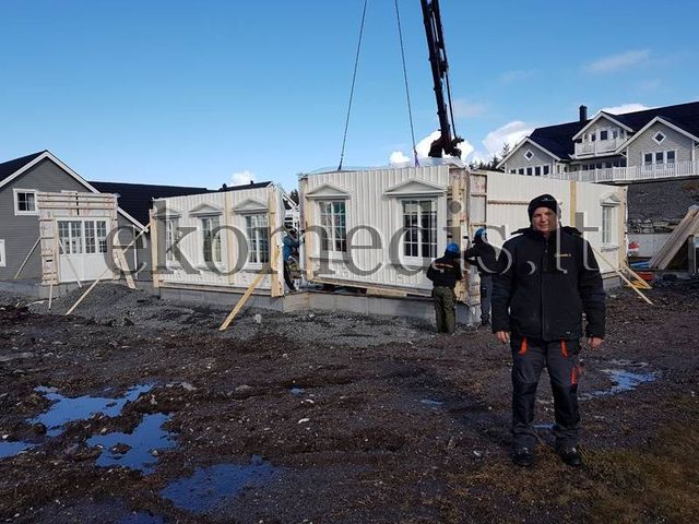Mann står foran elementhus under montering, der hvite veggelementer med vinduer heises på plass med kran